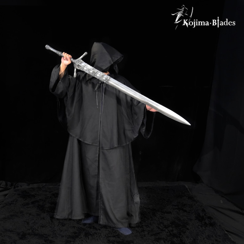 May include: A long, silver-colored metal sword with an ornate, textured crossguard and hilt, held horizontally. The sword features a pointed blade and intricate detailing on the guard. The Kojima Blades logo is visible in the top right corner.