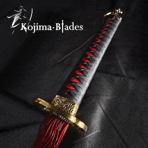 May include: A black and red katana sword with a gold-colored hilt and a red and black braided handle. The sword is on a black fabric background.