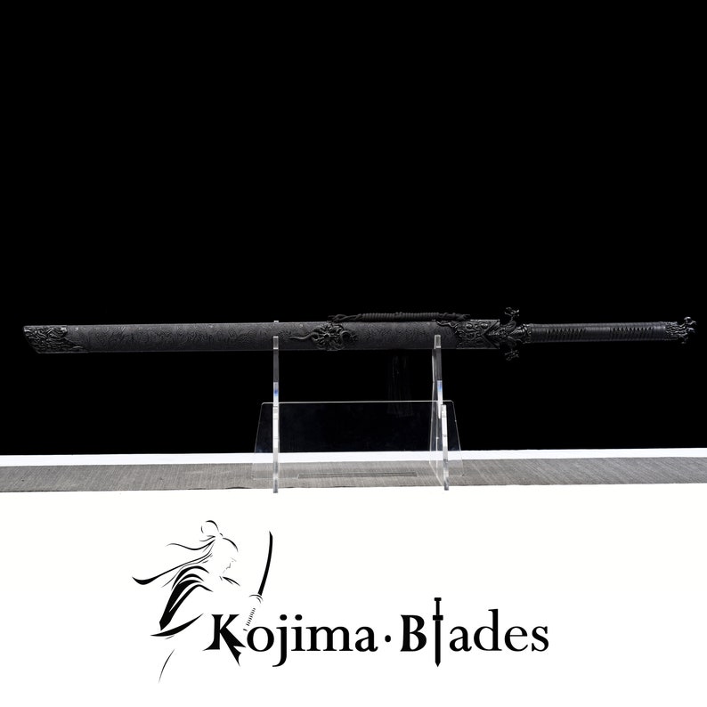 May include: A black katana sword with intricate dragon designs on the hilt and scabbard. The sword is displayed on a clear acrylic stand. The sword is labelled "Kojima Blades".