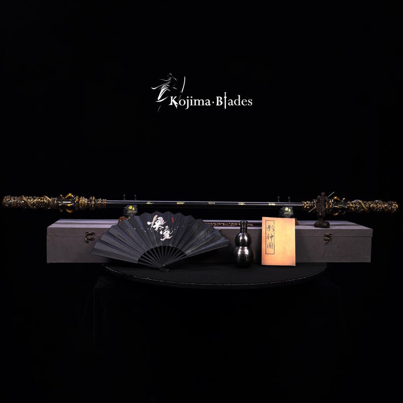 May include: A display of a long sword with ornate gold-colored dragon detailing, a black folding fan, a small metal gourd, and a book. The sword is displayed on a gray box. The text "Kojima Blades" is in the upper center.