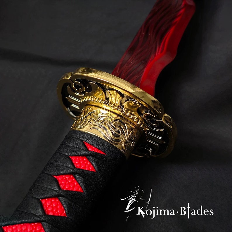May include: Close-up of a katana sword with a red blade and a gold-colored hilt. The handle is wrapped in black leather with red diamond-shaped accents. The sword features intricate gold detailing and the Kojima Blades logo.