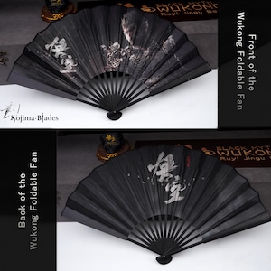 May include: Black foldable fan with a detailed illustration and Chinese characters. The fan is labeled "Wukong Foldable Fan" and features the "Kojima Blades" logo. The fan is made of black material and has a decorative design.