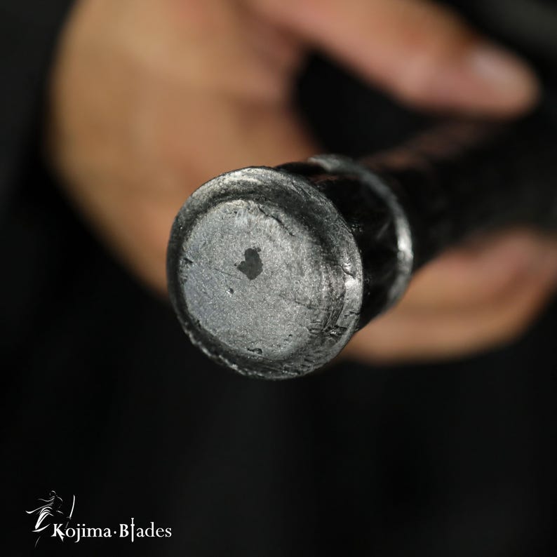 May include: A close-up view of the circular, textured metallic pommel at the end of a sword handle. The metal surface has a dark, weathered, and hammered appearance with a small, irregular dark spot in the center. The Kojima Blades logo is visible.