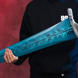 Op de afbeelding: Een blauw en zilveren zwaard met een gedetailleerd ontwerp op het lemmet. Het zwaard wordt vastgehouden door een persoon die een zwarte hoodie draagt.