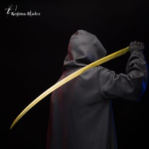 May include: A person wearing a black hooded robe holds a long, curved, gold-coloured sword with intricate engravings. The sword is held behind the person's back, with the tip pointing upwards.