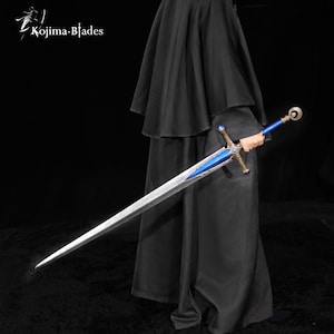 Op de afbeelding: Een lang zwaard met een metallic zilveren lemmet, een felblauwe pareerstang en een blauw handvat met versierde goudkleurige metalen accenten. Het zwaard heeft een ronde pommel en is voorzien van de tekst Kojima Blades in de hoek.