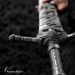 May include: A sword with a metallic, textured handle wrapped in dark, frayed fabric. The crossguard features a ridged, cylindrical design. The text Kojima Blades is visible in the bottom left corner, accompanied by a small sword icon.