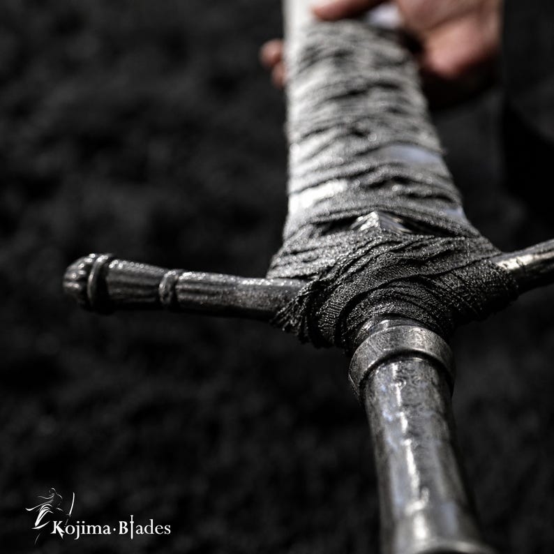 May include: A sword with a metallic, textured handle wrapped in dark, frayed fabric. The crossguard features a ridged, cylindrical design. The text Kojima Blades is visible in the bottom left corner, accompanied by a small sword icon.