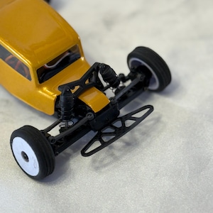 May include: A yellow toy car with black wheels and a black chassis. The car has a detailed front end with a grill and headlights. The car is on a white surface.