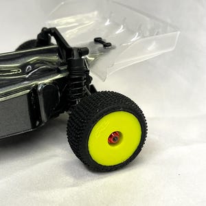 May include: A close-up of a black and grey RC car with a yellow wheel and a clear body. The car is on a white surface.