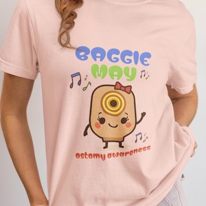 May include: A light pink t-shirt with the words "Baggie May" in blue and green, and the phrase "ostomy awareness" in orange. The shirt features a cartoon illustration of an ostomy bag with a red bow.