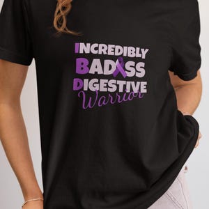 May include: Black t-shirt with the text "INCREDIBLY BADASS DIGESTIVE Warrior" in purple and white. The shirt features a purple awareness ribbon graphic. The shirt is a crew neck style.