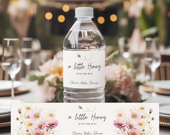 May include: Clear plastic water bottle with a white label that reads "a little Honey IS ON THE WAY Olivia's Baby Shower" with a bee graphic. A matching floral design is below the bottle. The bottle is on a table with floral arrangements.