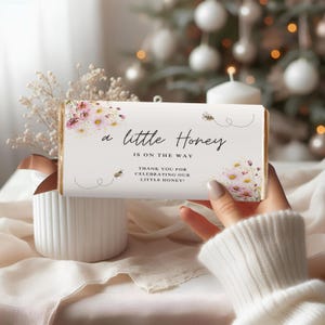 May include: A white candy bar wrapper with the text "a little Honey IS ON THE WAY" and floral and bee illustrations. The wrapper also says "THANK YOU FOR CELEBRATING OUR LITTLE HONEY!" The wrapper is held in front of a blurred background.