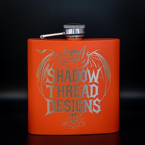 Op de afbeelding: Oranje roestvrijstalen heupfles met een zilveren dop en scharnierende tuit. De fles heeft een zilveren afbeelding van een schedel met vleermuisvleugels en de tekst "SHADOW THREAD DESIGNS".