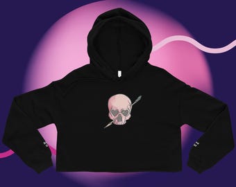 May include: Black cropped hoodie with a hood and long sleeves. The front features a pink skull with heart-shaped eye sockets pierced by an arrow. The background is a gradient of pink and purple.