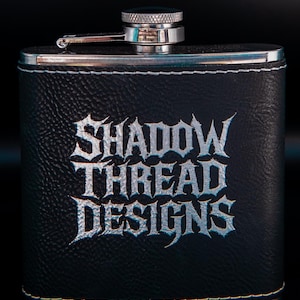 Könnte beinhalten: Schwarze Lederflasche mit dem Text "SHADOW THREAD DESIGNS" in silbernen Buchstaben. Die Flasche hat einen silbernen Verschluss und einen kleinen Klappdeckel. Weiße Nähte umranden die Kanten.