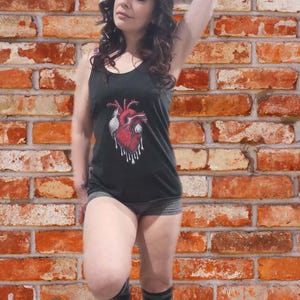 May include: A woman wearing a black tank top with a red anatomical heart graphic. The tank top has a scoop neck and is sleeveless. The woman is standing in front of a brick wall.