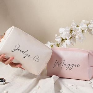 Puede incluir: Dos bolsas de maquillaje personalizadas, una blanca con el nombre "Jocelyn B" y otra rosa con el nombre "Maggie".