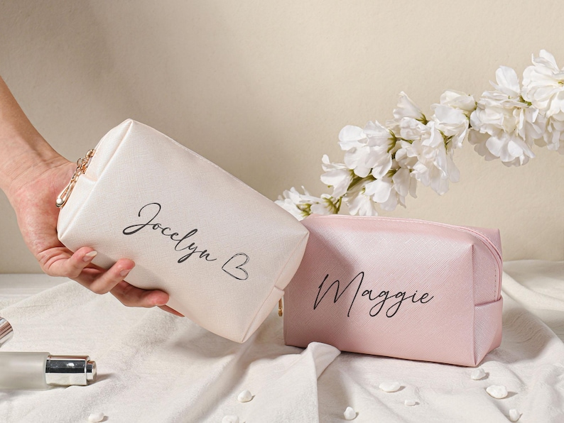 Puede incluir: Dos bolsas de maquillaje personalizadas, una blanca con el nombre "Jocelyn B" y otra rosa con el nombre "Maggie".