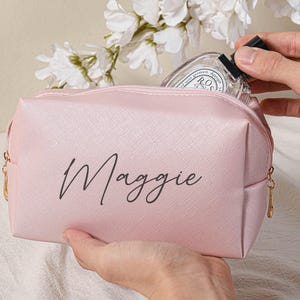 Puede incluir: Una bolsa de maquillaje rosa con cremallera y el nombre "Maggie" impreso en letra negra en la parte delantera.