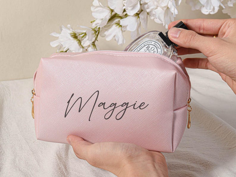Puede incluir: Una bolsa de maquillaje rosa con cremallera y el nombre "Maggie" impreso en letra negra en la parte delantera.