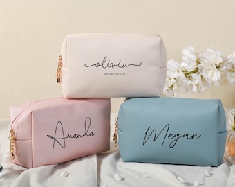 May include: Three personalized cosmetic bags with gold zipper pulls. The bags are pink, white, and blue. The names "Amanda", "Olivia Bridesmaid", and "Megan" are printed on the bags in a script font.