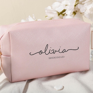Puede incluir: Una bolsa de maquillaje rosa con cremallera con el nombre "Olivia" y la palabra "bridesmaid" impresa en ella.