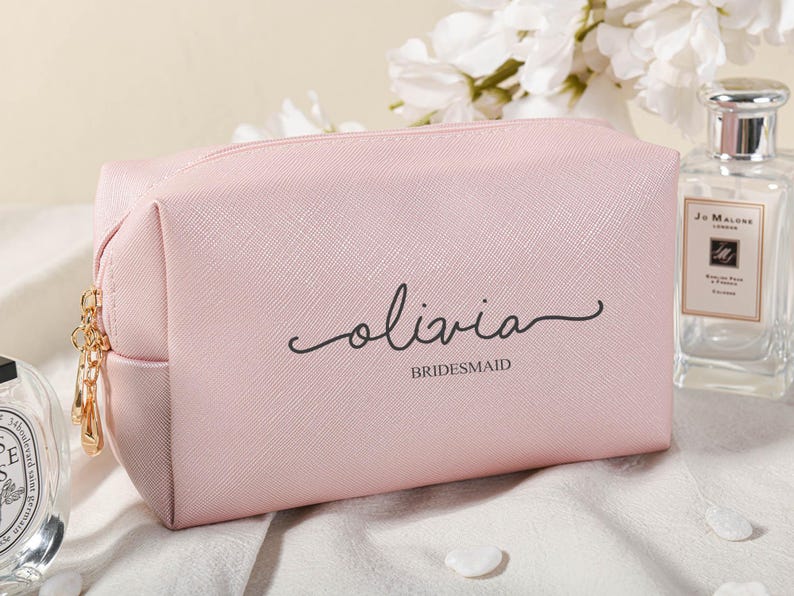 Puede incluir: Una bolsa de maquillaje rosa con cremallera con el nombre "Olivia" y la palabra "bridesmaid" impresa en ella.