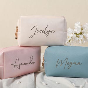 Puede incluir: Tres bolsas de cosm&eacute;ticos personalizadas con cierres de cremallera. Las bolsas son de color rosa, blanco y azul con los nombres Amanda, Jocelyn y Megan escritos en cursiva negra.
