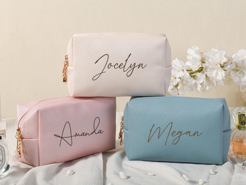 Puede incluir: Tres bolsas de cosm&eacute;ticos personalizadas con cierres de cremallera. Las bolsas son de color rosa, blanco y azul con los nombres Amanda, Jocelyn y Megan escritos en cursiva negra.