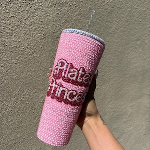 May include: Pink tumbler covered in rhinestones with the words "Pilates Princess" in a contrasting red and white font. The tumbler has a clear straw and a rhinestone-studded rim.