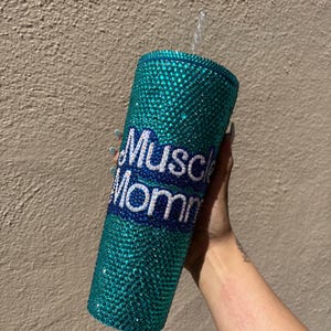 Puede incluir: Un vaso de color turquesa adornado con pedrería y una pajita. El vaso tiene las palabras "Muscle Mom" en letras blancas y azules. El vaso está cubierto de pequeños y brillantes pedrería.