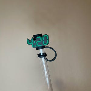 Puede incluir: Una botella de agua cilíndrica transparente con una tapa negra y una pantalla digital verde que muestra "4:20". La botella tiene un lazo negro adjunto a la tapa. El fondo es de un color neutro sólido.