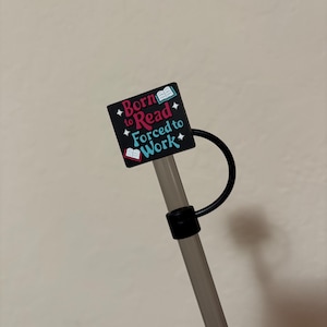 May include: A black and grey straw with a black square charm that reads "Born to Read Forced to Work" in red, blue, and white. The charm has a book graphic on each side. The straw has a black ring and a black handle.