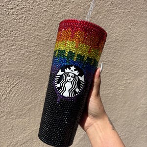 Op de afbeelding: Een met glinsterende kristallen versierde Starbucks-beker met een regenboogverloop. De kleuren lopen van rood aan de bovenkant, via geel, groen en blauw, naar een zwarte basis. Het Starbucks-logo is zichtbaar.