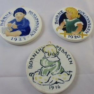 Peut inclure: Trois ornements ronds en céramique avec un fond blanc et des motifs peints bleus et jaunes. Chaque ornement représente un enfant dans une pose différente et le texte "Børne Hjælpsdagen" et une année : 1916, 1920 et 1927.