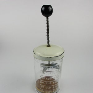 Peut inclure: Un hachoir à aliments en verre vintage avec un couvercle blanc et une poignée noire. Le bocal en verre comporte des graduations en tasses et en onces. Le hachoir contient un aliment brun.