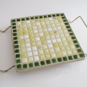 May include: A square ceramic hot pad with a green and yellow mosaic design. The hot pad has a metal frame with two handles.