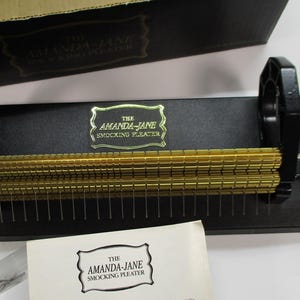 May include: The Amanda-Jane Smocking Pleater is a black sewing tool with two horizontal gold-coloured metal rollers featuring fine grooves and a series of thin needles. It includes a black base, a side adjustment knob, and a branded label.