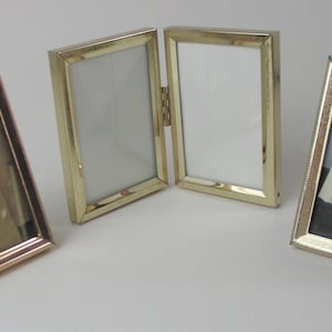 May include: Four gold-coloured picture frames in different styles. One is a single frame, another is a double-hinged frame, and the others are single frames with a stand. The frames have a textured border.