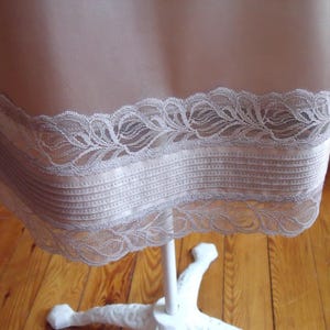 May include: A beige slip with a delicate lace trim. The lace is a light grey colour and features a floral design. The slip is made of a smooth, silky material and is displayed on a white stand.