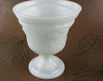 May include: White milk glass vase with an embossed grape design. The vase has a wide mouth and a round base.
