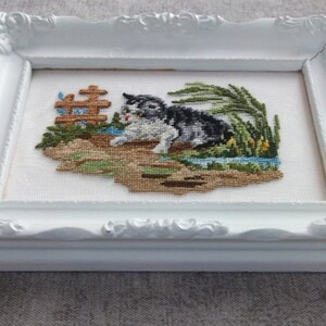 May include: A white frame with a white mat surrounding a cross-stitch of a black and white cat in a green grassy area. The cat is looking to the left and has its mouth open. There is a small brown wooden fence in the background.