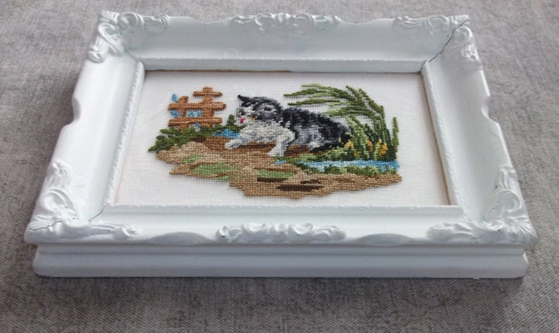 May include: A white frame with a white mat surrounding a cross-stitch of a black and white cat in a green grassy area. The cat is looking to the left and has its mouth open. There is a small brown wooden fence in the background.