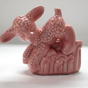 May include: A pink ceramic lamb figurine with a textured body and a reclining pose. The lamb is resting on a decorative base. The figurine has a glossy finish and is likely a decorative item.