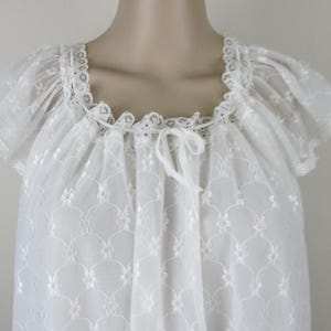 May include: A white nightdress with a square neckline and short, frilled sleeves. The nightdress has floral embroidery and lace trim on the neckline and sleeves. A white ribbon tie is at the neckline. The fabric is semi-sheer.