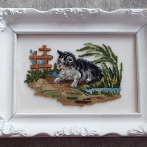 May include: A white framed cross-stitch embroidery of a black and white cat sitting in a grassy area with a small wooden fence and blue flowers.