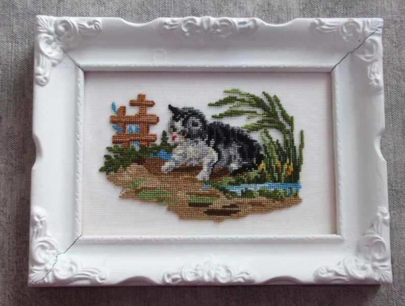 May include: A white framed cross-stitch embroidery of a black and white cat sitting in a grassy area with a small wooden fence and blue flowers.