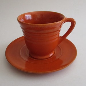 Peut inclure: Une tasse à thé et une soucoupe en céramique orange vif. La tasse a une forme arrondie avec un bord évasé et une anse. La soucoupe a une forme simple et circulaire.